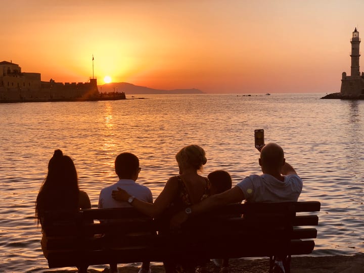 People silhouetted on a bench watch a fiery sunset over the sea, a lighthouse on the far mole.