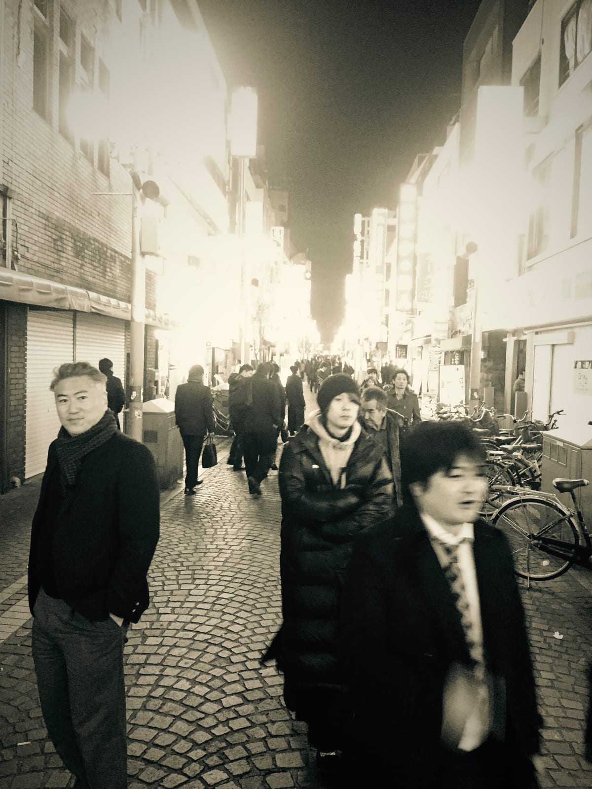 A black-and-white street scene, two figures walking toward the camera among bicycles.