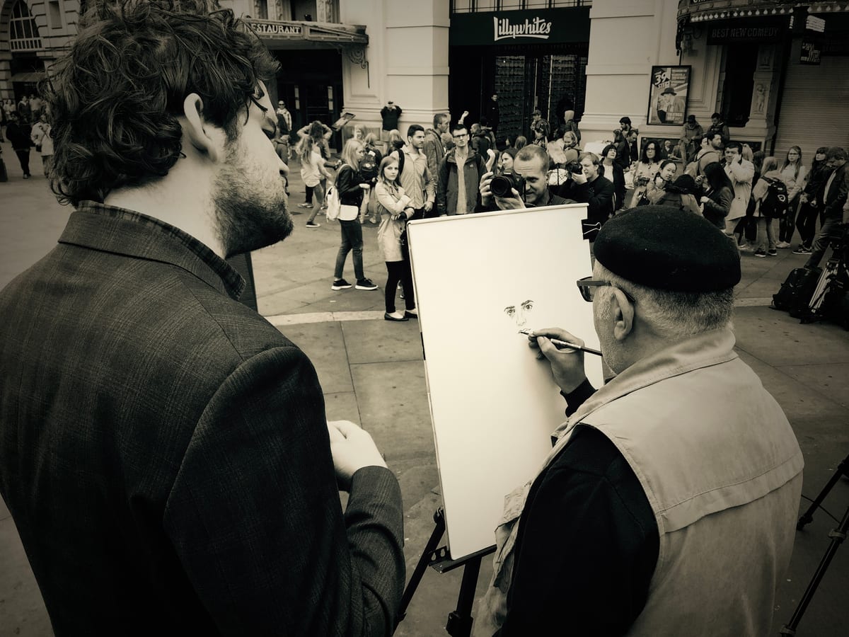 In black and white, a street artist in a beret sketches a young man who poses, a crowd behind.